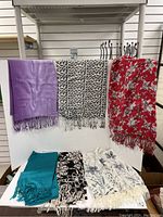 Photo of six scarves displayed on a white background, three hanging and three folded, showing a variety of colors and patterns including purple, black and white spots, red floral, teal, black floral, and white floral.