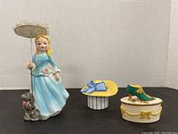 Porcelain girl figurine with dog holding a parasol umbrella next to small decorative shoe and hat boxes.