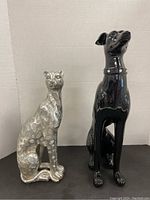 Black ceramic dog statue and silver composite cat statue side by side against a neutral background.