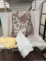 Various tablecloths and runners displayed including the large white Laura Ashley tablecloth partially hanging and several folded linens on a white board and chair.