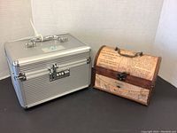 Two boxes side by side: silver metal box with combination lock and a decorative wood box with newspaper print covering, both placed on black surface.