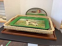 Three stacked trays shown; largest is wooden, middle is white wicker, and smallest is green tin tray with dog painting