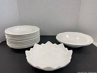 Stack of 12 medium white plates with scalloped edges and 2 large white bowls with scalloped edges behind, displayed on black surface against gray background.