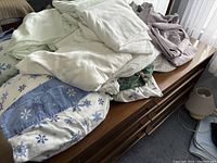 Photo showing six sets of folded bedding including fitted sheets, flat sheets, and pillowcases in various colors and patterns including blue with snowflakes, green floral, white, and light gray.