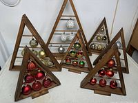 Five triangular wooden displays with horizontal rods holding various Christmas ball ornaments in red, gold, white, silver, green, and multicolor.