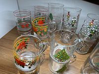 Top-down angled photo showing a collection of Christmas-themed glass mugs and glasses with designs including poinsettias, Christmas trees, nutcracker figures, and snowflakes on a wooden surface.