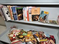 Multiple spiral-bound and paperback cookbooks displayed on shelves, some vintage titles visible with illustrated covers showing food dishes.