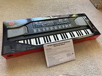 Electric keyboard piano in black box with visible keys and two built-in speakers, with operating manual placed in front.