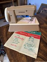 Front-right angle view of miniature Singer sewing machine with beige and cream color, hand crank visible. Includes original paper manual showing a drawn girl and machine.