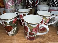Close-up of four coffee mugs with red rose floral patterns and gold trim on rim and handles.