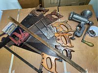 Photo showing multiple vintage wooden-handled hand saws with different handle designs, heavy duty Dominion meat grinder, red staple gun, metal ruler.