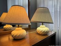 Pair of ceramic table lamps on a wooden table, showing the cream swirl design bases and beige pleated fabric shades, with one lamp lit and the other off.