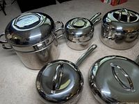 Complete set of 8 polished stainless steel cookware pieces with fitted lids arranged on floor, showing variety of sizes and shapes including stockpots, saucepans, and skillets.