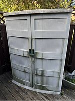 Front view of closed gray Suncast plastic shed showing two doors with central locking handle and visible surface dirt and weathering.
