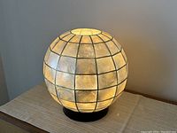 Front side view of globe-shaped mother of pearl table lamp illuminated, showing grid pattern of square panels and black base.