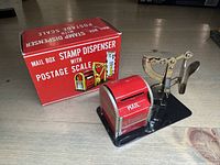 Red mail box style stamp dispenser and metal postage scale on black base, original red box behind them.