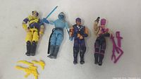 Four G.I. Joe action figures posed laying flat against a white background, showing full body and accessories beside them including yellow weapons and a blue sword.