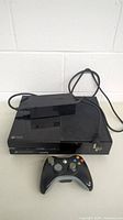 Xbox One console with controller and power cord placed on table, front view showing entire set.