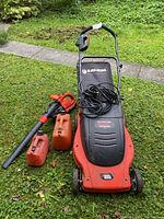 Black & Decker electric lawn mower with attached grass bag, power cord, gas leaf blower, and two red fuel containers placed on grass.