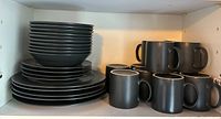 Black plates stacked with bowls and black mugs on right side in cupboard shelf.