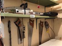 Shelf containing electric chain saw on top with Oregon OE guide bar, and three handheld saws hanging below with wooden handles and metal blades.