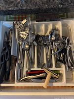 Top down view of kitchen drawer showing stainless steel cutlery grouped by type with knives, forks, spoons, plus various kitchen utensils and tools including scissors and corkscrews organized in tray compartments.
