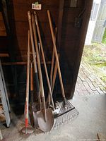 Photo of the garden tools leaning against a dark wooden wall showing two shovels, a hoe, and two rakes with wooden handles.