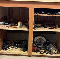 Tools stored in wooden cabinet lower shelf including power tools with cords such as circular saw and angle grinder.