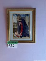 Front view of framed needlepoint artwork showing Virgin Mary and baby Jesus, with green tape indicating dimensions (13"w x 16"h) and lot number.