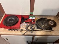 Multiple saw blades laid out on a counter including one large carbide-tipped circular saw blade in a red case, several stacked blades including abrasive cutting discs, and two C-clamps