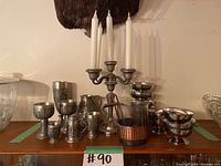 Wide view of the full lot showing pewter cups, mugs, bowls, and three-arm candelabra with white candles arranged on wooden surface