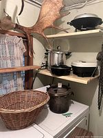 Shelf with multiple kitchen appliances including stainless steel pressure cooker, cast iron pot, electric skillet and large woven basket on surface.
