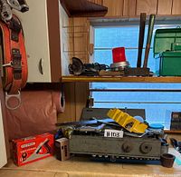 Metal toolbox with three compartments filled with assorted hand tools including screwdrivers, pliers, drill bits and a socket set on a blue holder, placed on countertop beside a red tool belt hanging and a planer tool visible.