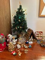 Small decorated green artificial Christmas tree with vintage candle lights, wooden nativity scene, assorted figurines and boxed Christmas ornaments spread around a wooden table.
