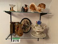 Full view of the lot showing porcelain vase with floral and gold detailing, porcelain dog figurine, small bronze pitcher, framed embroidery art, wooden carved piece, and small statues on glass shelves (not included).