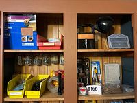 Wooden cupboard shelf with assorted tools including jars with screws, yellow bins with drill bits, tape rolls, battery, flashlight, and lamp.