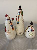 Image showing three ceramic snowmen figurines of different heights (7, 10, and 12 inches) with multicolored scarves, black and red hats, and snowflake decorations on their bodies.
