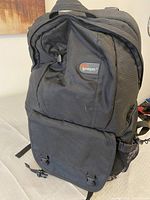 Front view of black Lowpro camera backpack showing logo patch, front zippered and buckle flap compartments, and overall design.