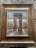 Photo of an oil painting depicting a Parisian street scene centered on a large stone arch with people walking along a tree-lined pathway. The frame is wooden and distressed.