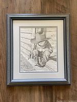 Framed monochromatic print depicting a child with a large hat sitting against a log cabin wall, signed by artist Bernie Brown