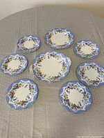 Top view of all 8 pieces arranged on table showing matching floral and blue border designs, 7 smaller plates and 1 larger handled plate.