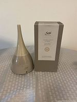 Photo of beige Saje Aroma Genie ultrasonic diffuser next to its box on a fabric surface.