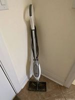 Full view of Shark steam mop upright against wall, showing white and gray body and rectangular mop head without cloth.