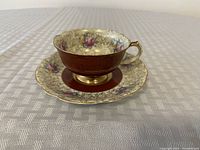 Porcelain teacup sitting inside matching saucer showing gold filigree pattern, pink roses, and reddish-brown band with gold accents