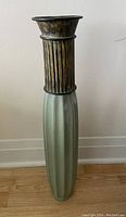 Tall decorative vase with metallic ribbed top portion and green elongated base against plain wall.