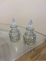 Pair of vintage glass perfume bottles with ornate geometric cut glass patterns and clear plastic diamond-shaped stoppers on top, displayed on glass surface in front of beige wall.