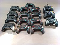 Top-down view of all 16 controllers spread out on table, showing 4 black wireless Xbox Series X controllers, 9 black generic wireless controllers with colored accent lines, and 3 black wired Xbox 360 controllers with colored buttons.