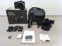 Full lot with digital camera, box, tripod, carrying case, cables, batteries, memory card, strap, and manual displayed on table.