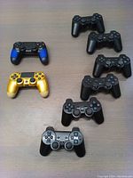 Top-down overview showing all 8 controllers arranged on table. Includes 6 black PS3 controllers and 2 distinct PS4 generic controllers: one gold with black trim, one black with blue grips.