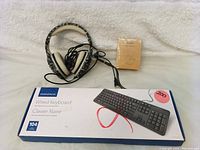 Insignia wired keyboard box shown alongside a wired headset and other item on a white textured surface for scale and condition assessment